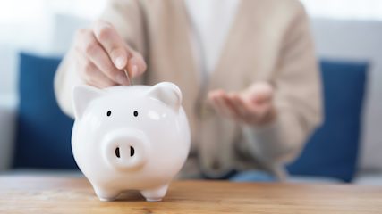 A person wearing a white shirt and beige cardigan putting coins into a white piggy bank