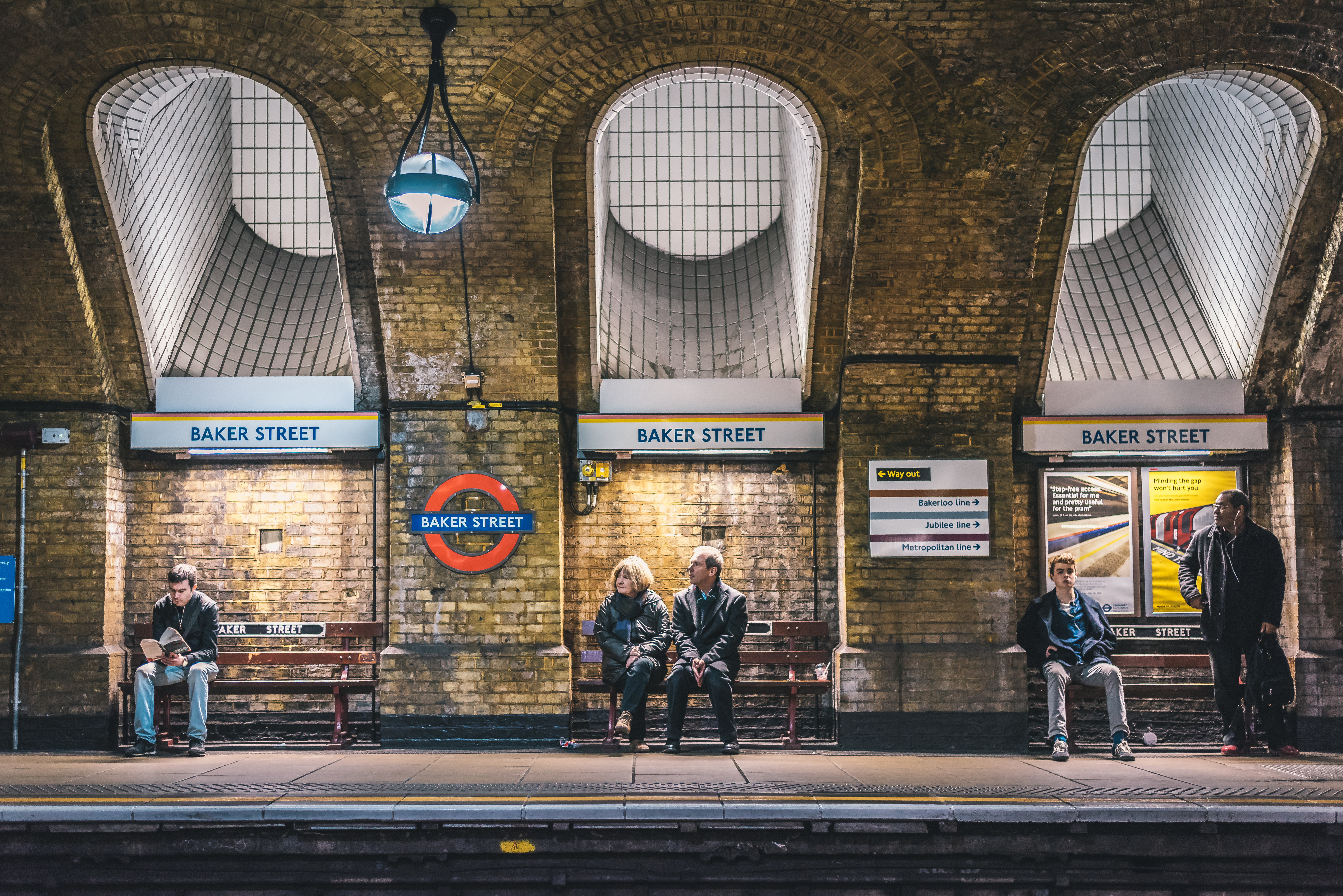 Baker Street Underground platform with ‘Baker Street’ station signs, roundel logos and passengers waiting on the platform.