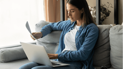 Young woman sitting on a sofa with a laptop and paperwork in front of her.