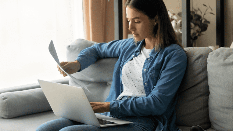 Young woman sitting on a sofa with a laptop and paperwork in front of her.