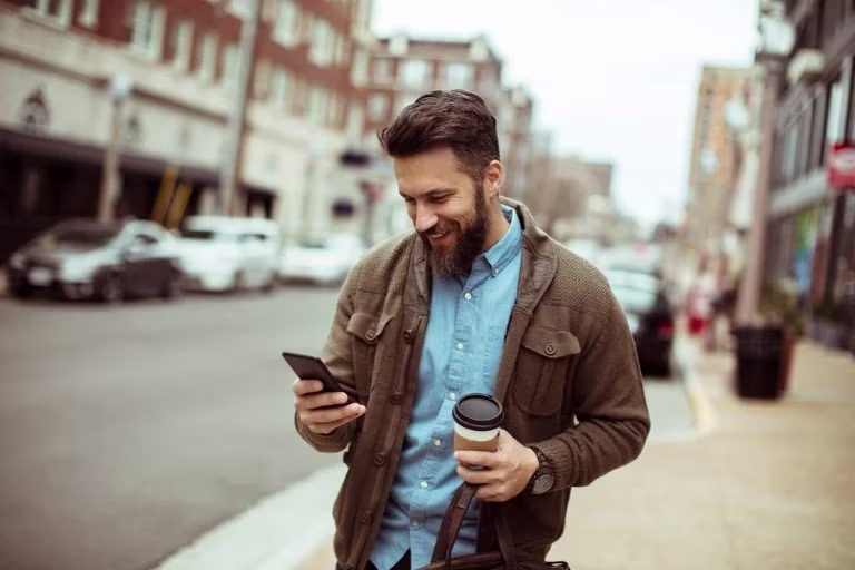 A bearded brunette Caucasian man looks at his mobile while walking down the street.