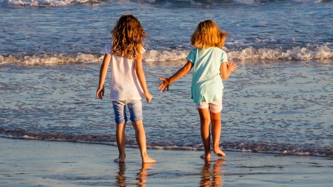 Image shows two young children on a beach, playing in the shallows of the sea. They're around 6 or 7 years old, wearing shorts and t-shirts and have their backs to the camera. The sunlight implies it's close to the end of the day.