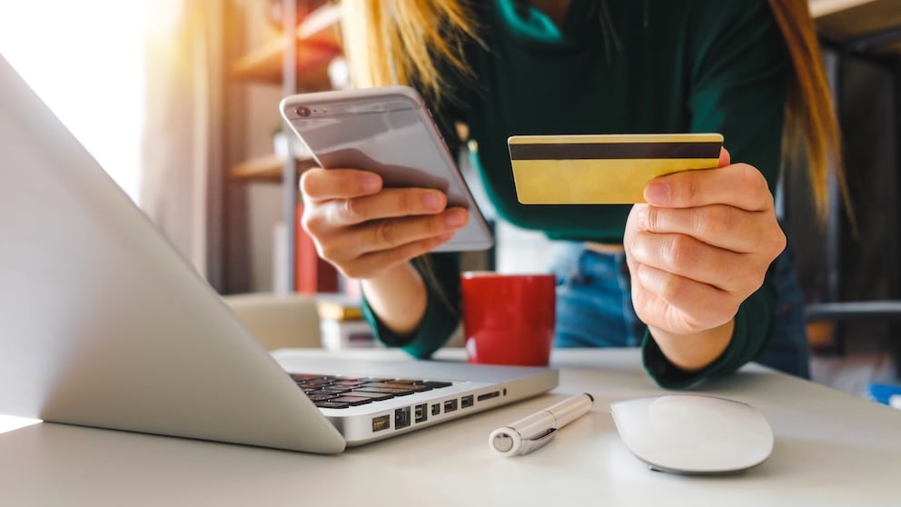 Women in front of a laptop holding a smart phone in one hand and a payment card in the other.