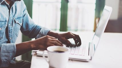 Close-up photo of someone's hands typing on a laptop with a cup of coffee in the foreground.