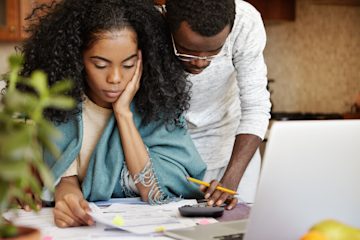 Heterosexual couple, likely in their thirties, studying documents on a table and using a calculator. The woman looks stressed.