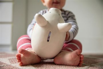A baby wearing pink and white stripes, holding a white piggy bank