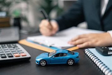 A blue toy sports car on a desk next to a calculator and a person writing on a clipboard