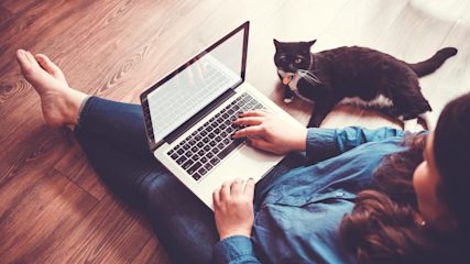 Woman sitting on a wooden floor using a laptop with a black and white cat laying nearby.