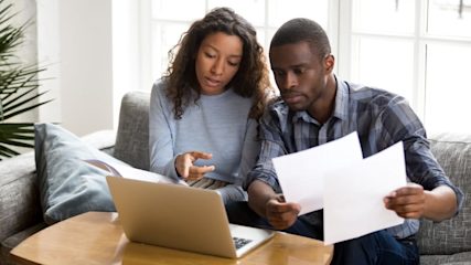 A woman and a man looking at a laptop together while the man holds several papers.