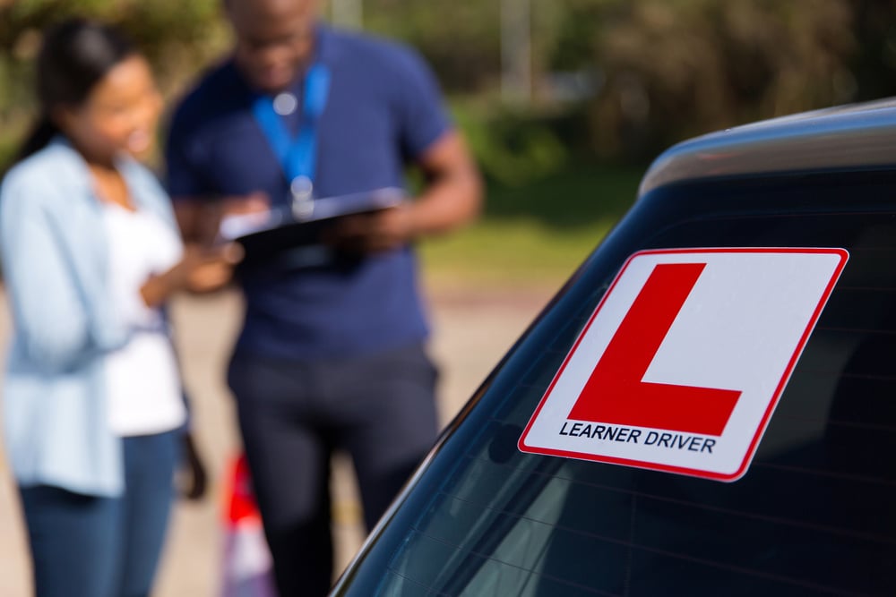 Close-up of a badge on the back of a car, featuring a large red L and the words "learner driver", with two people out of focus in the background.