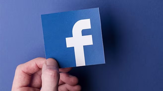 Close up photo of a left hand holding a cut out of the Facebook logo over a blue surface.