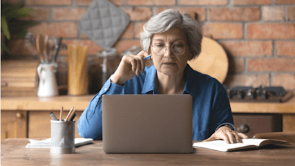 An older woman sat at a kitchen table using a laptop.