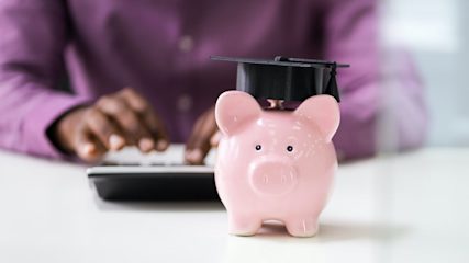 A pink piggy bank with a graduation cap sitting on a table in front of a person typing on a calculator.