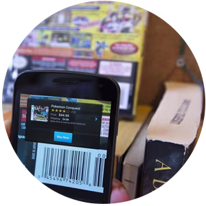 A smartphone with the Amazon app being used to scan a barcode and displaying product details for Pokémon Conquest, including price and purchase options.