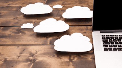 Close-up photo of cut-outs of white clouds on a wooden table top with a silver and black laptop to the right.