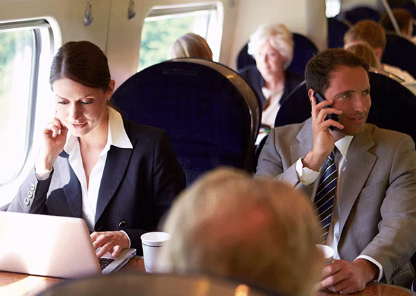 Two commuters sitting at a table on a train, one using a laptop and the other making phone calls.
