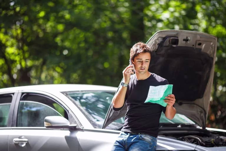 Young man leaning on his car. He's on his mobile with a document in his hand and the bonnet of the car is up.