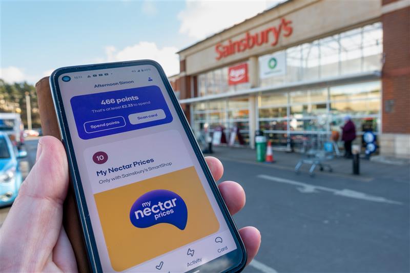 Shopper holding a supermarket basket filled with groceries and a smartphone showing the Nectar app