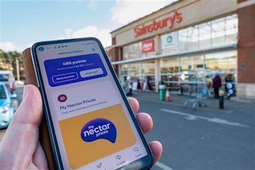 Shopper holding a supermarket basket filled with groceries and a smartphone showing the Nectar app