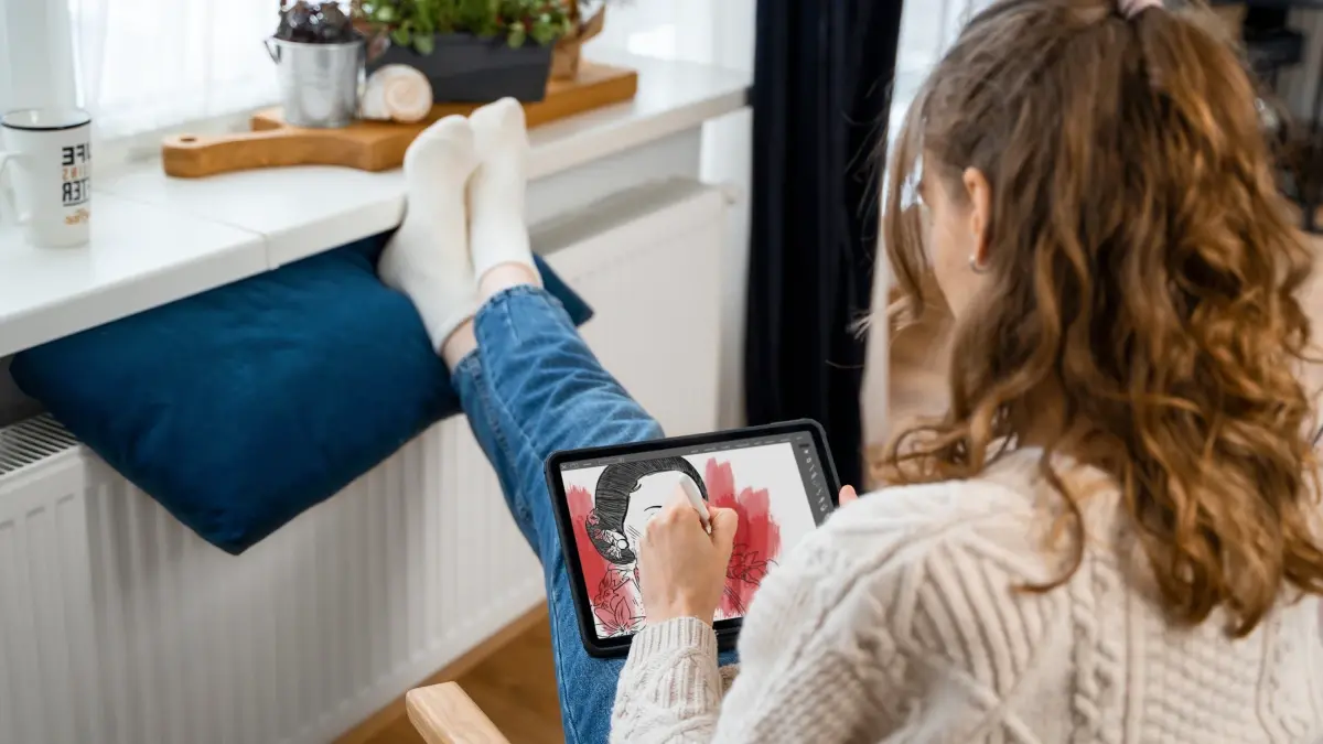 Foto van jonge dame met sokken aan die met voeten op radiator zit. Ze tekent op een tablet.