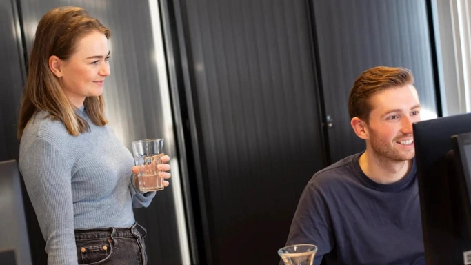 2 collega's op kantoor, een vrouw met een glas water in haar hand en een man aan de pc