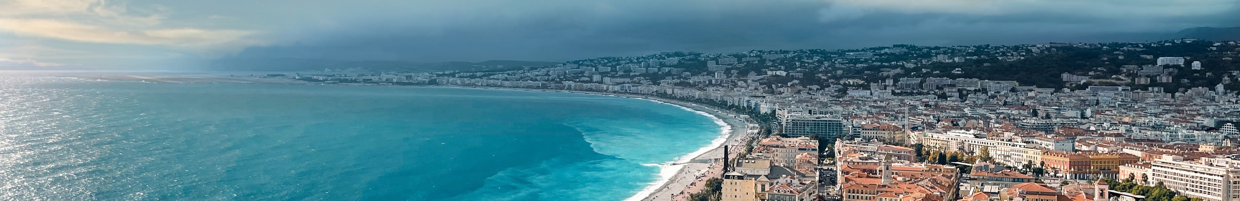 Vue aérienne de la Promenade des Anglais à Nice, bordée par une mer bleue, avec des nuages à l’horizon.
