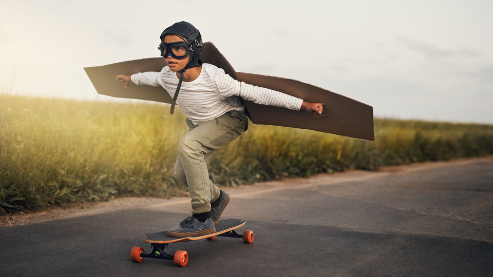 A child on a skateboard with cardboard airplane wings and flight goggles and a helmet. Riding down a paved road. 