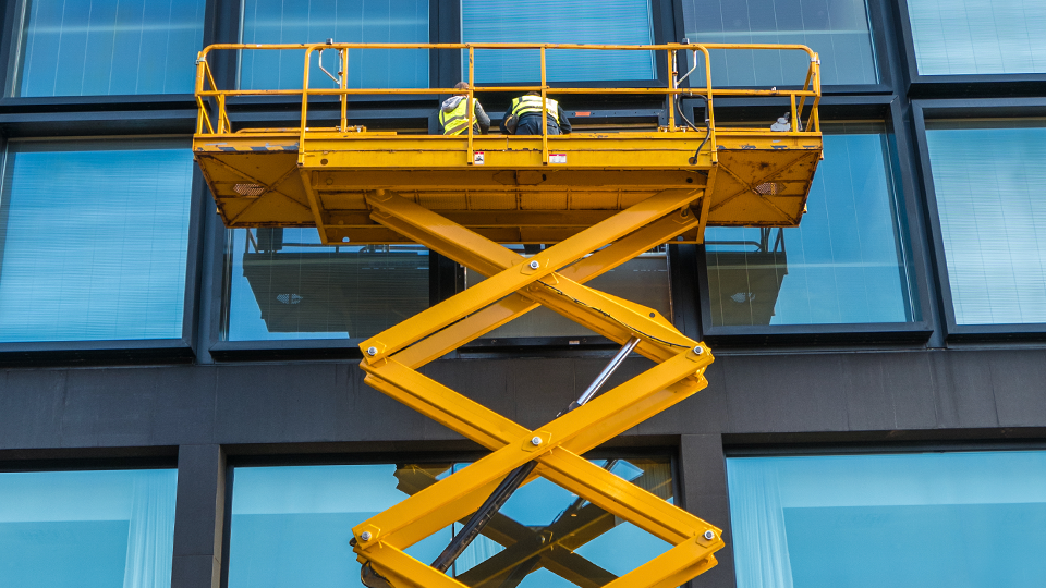 A photo of a scissor lift hoisting up two maintenance workers up the side of a tall glass building.