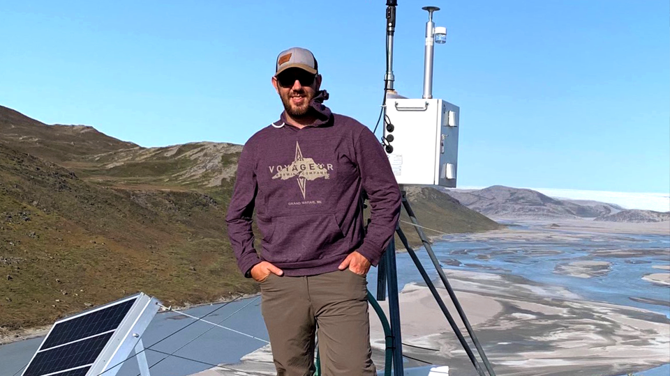 Man in sunglasses, hat, and sweatshirt standing in front of a mountainous landscape. 