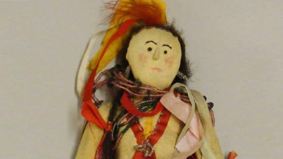 Standing male doll wearing tan hide suit, leggings, and moccasins with pink and red diamond design ribbonwork. Dark hair woven in two braids with red ribbons and feathers. Glass bead necklace and earrings with cotton thread. Bow in one hand, arrow in the other, and quiver on back.