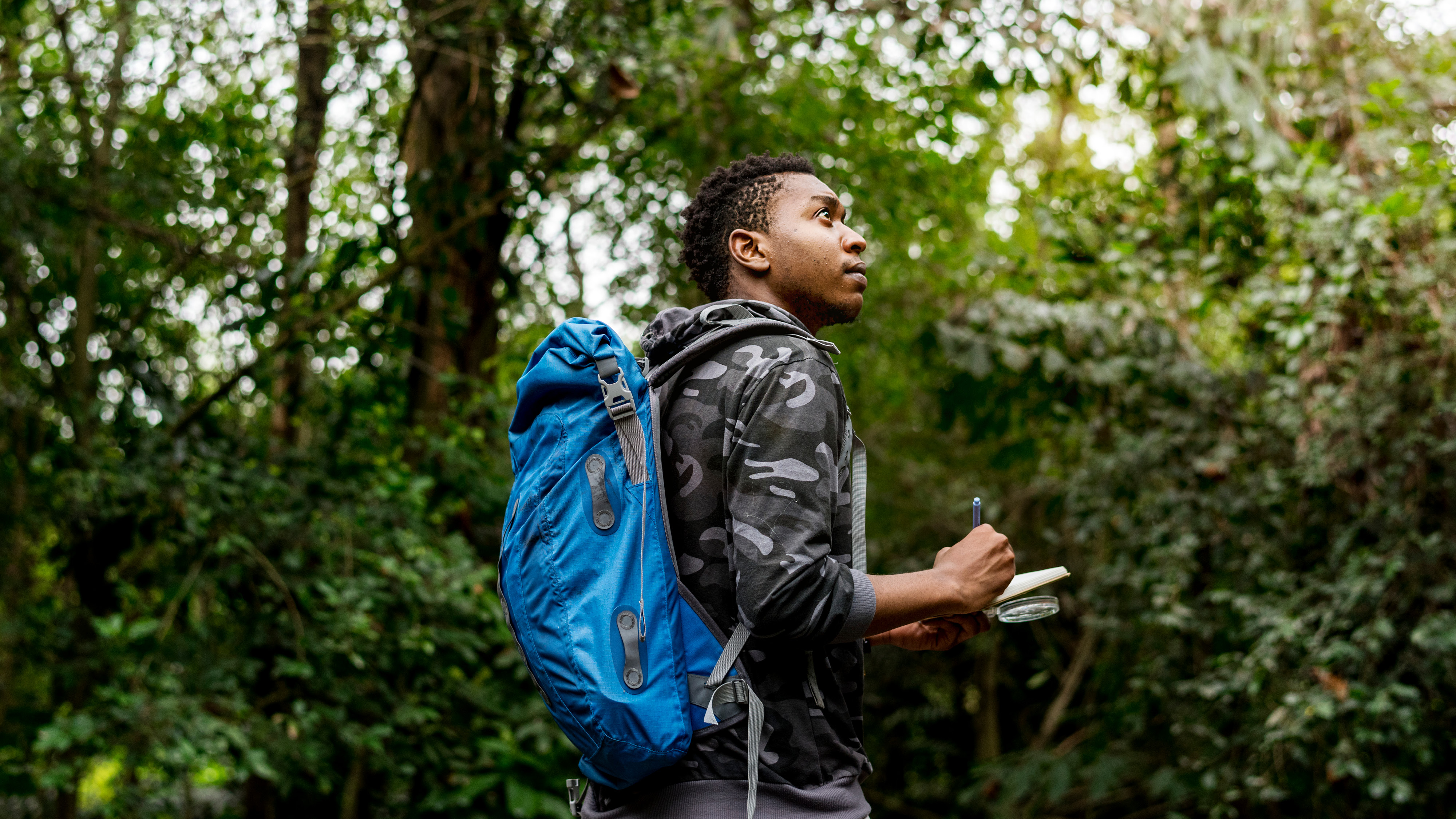 A person hiking in the woods with a backpack, stopping to take a note in a notebook. 