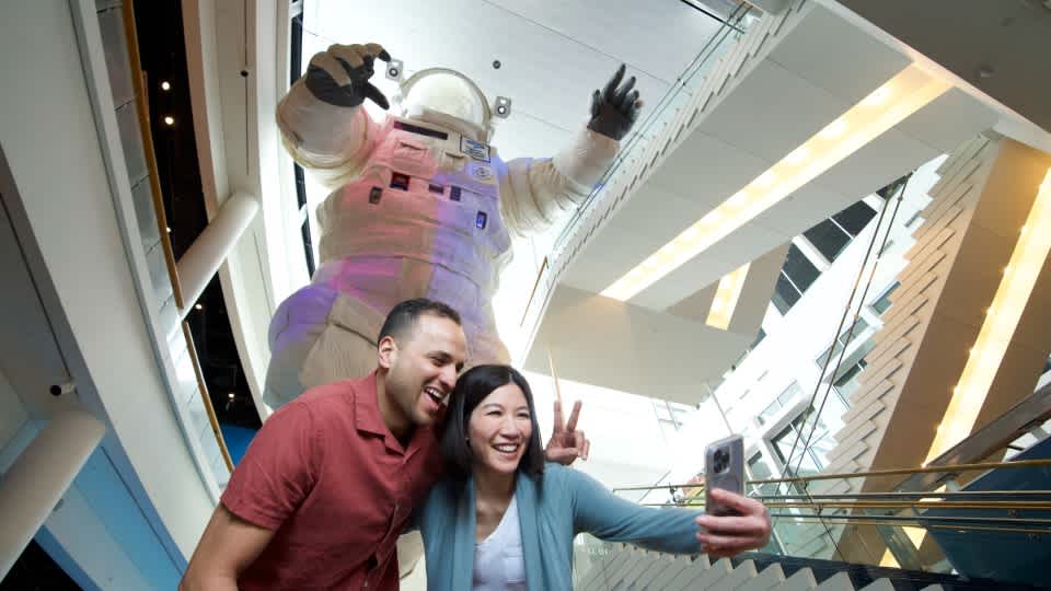 Two adults posing for a selfie in front of the giant astronaut. 