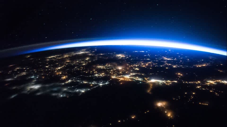 A photo of Earth from space, only a small portion of Earth is visible. It's night - city lights on Earth can be seen with a blue horizon emerging behind Earth.