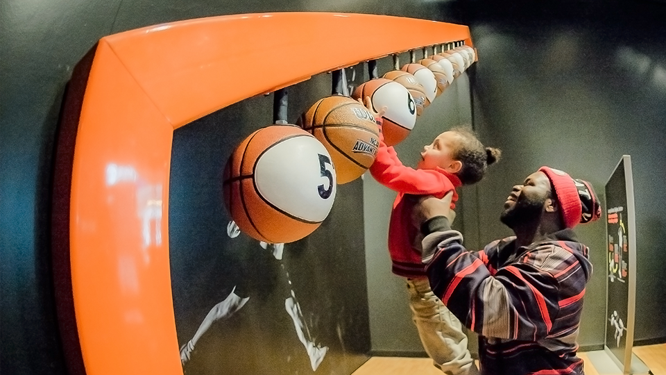 An adult holding a child up to touch a basketball in the Sportsology exhibit