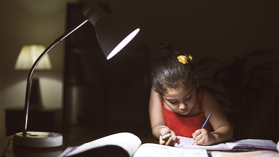 A young girl doing homework at a desk by lamplight. 