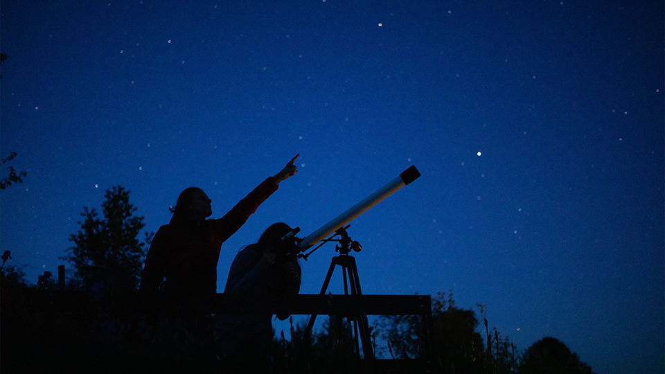 A pair exploring the night sky - one glancing through a telescope while the other points to the sky.