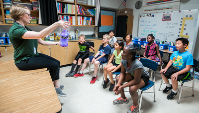 Photo of a classroom. A museum staff member demonstrated a science experiment in front of children.