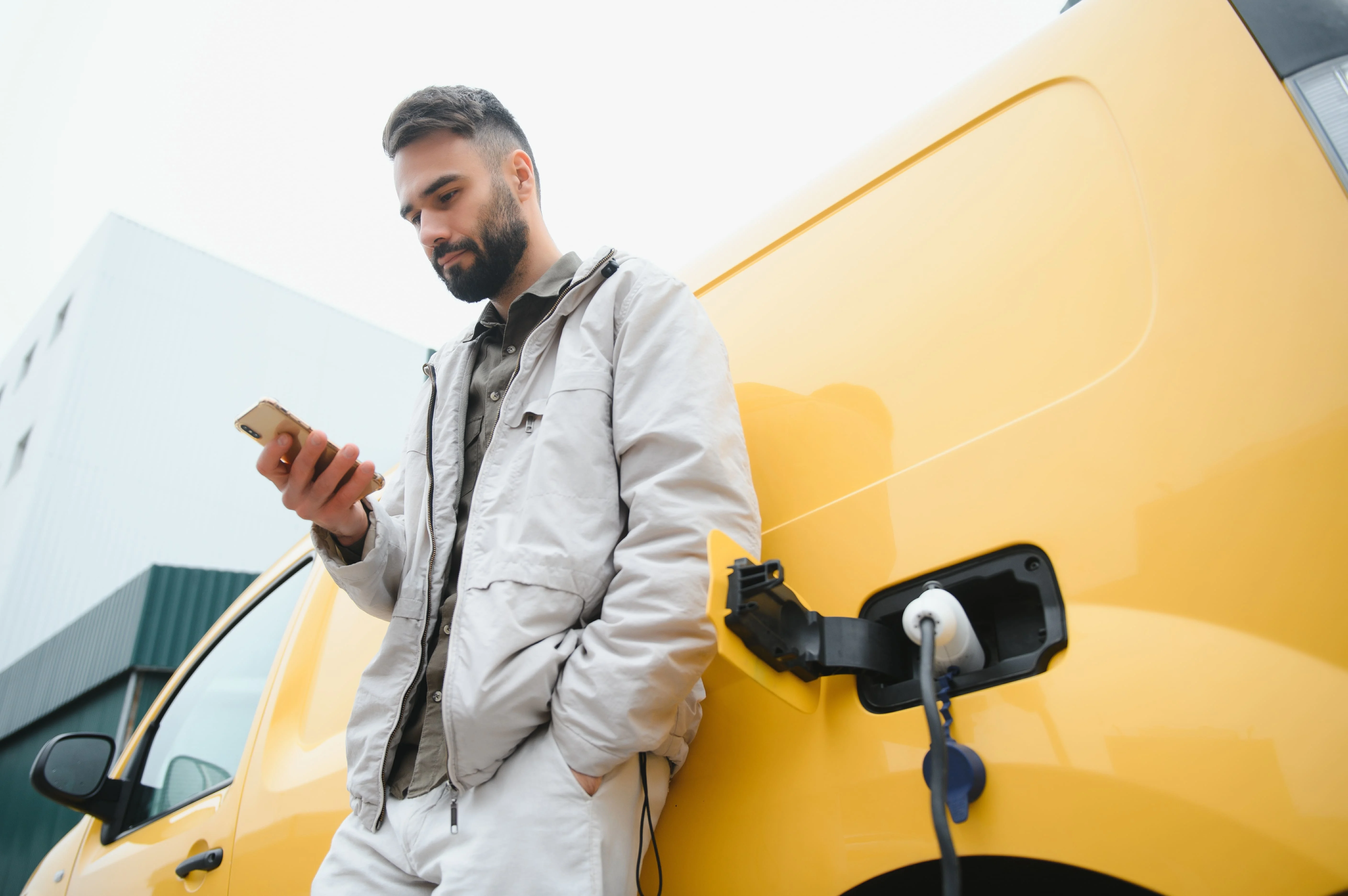 Un homme adossé à une voiture électrique jaune utilise son smartphone pendant que la voiture se recharge. 