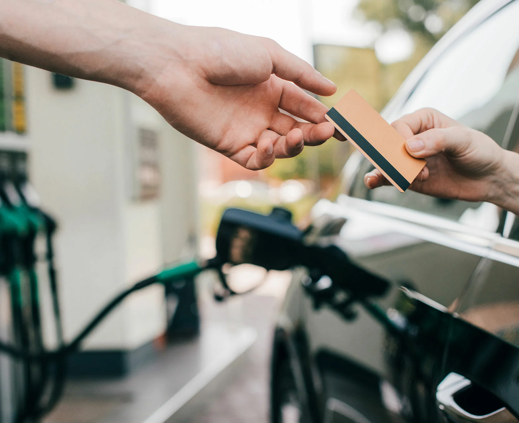 Eine Person reicht einer anderen eine Tankkarte vor dem Hintergrund einer Tankstelle mit Zapfsäulen.