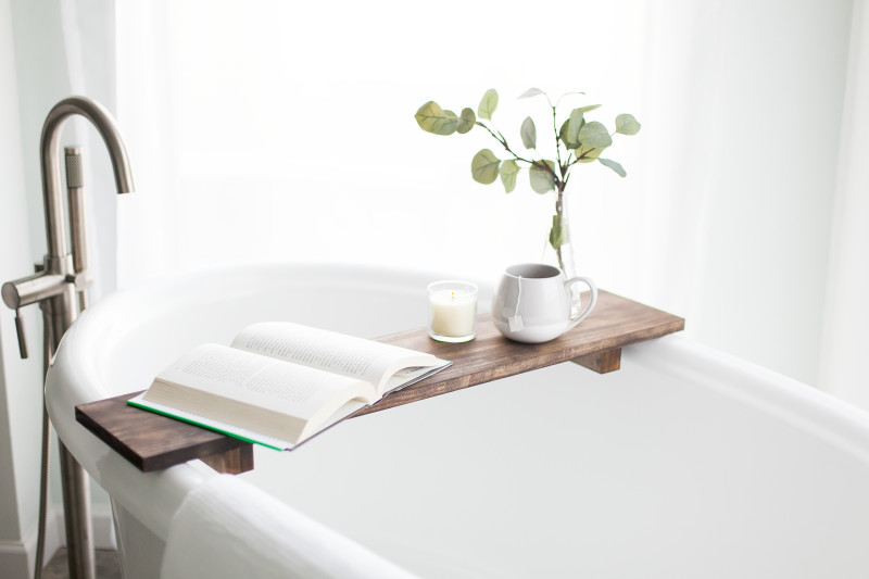 A closeup of a white oval bathtub with a freestanding faucet  and a wooden bathtub tray holding a book, candle, cup of tea and plant.