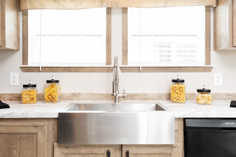 Manufactured home kitchen featuring a stainless steel farmhouse sink, marble style counters and light wood cabinets with matching light wood trim.