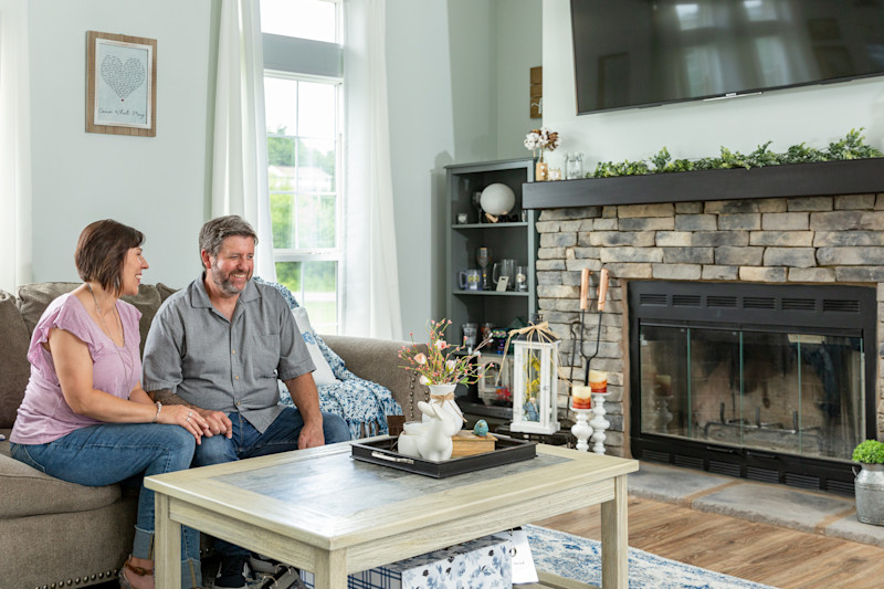 Woman and man sit on brown couch in the living room of a manufactured home with coffee table in front of them next to a stone fireplace with a TV above it, and a bookshelf and window behind them.