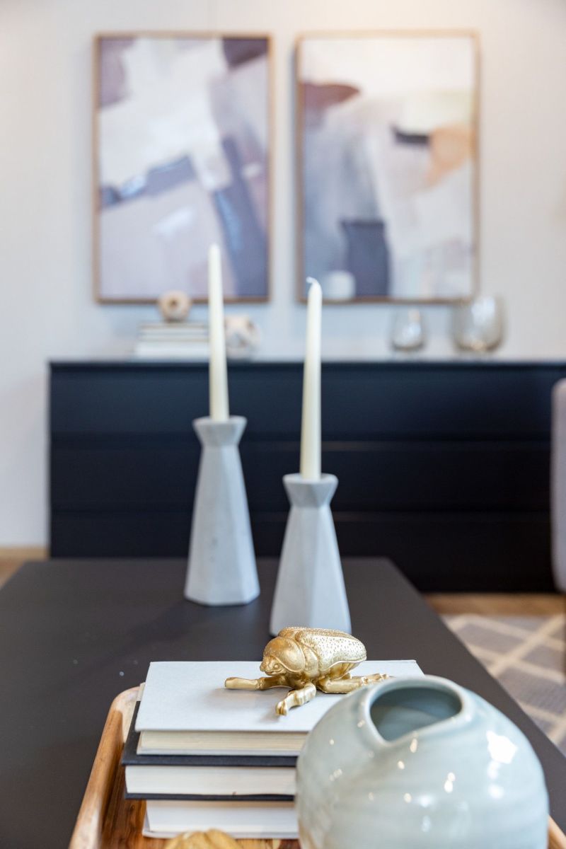 Two abstract art pieces are hung in the background with a black dresser below them. Close up there are two gray candlesticks with some décor on a coffee table.