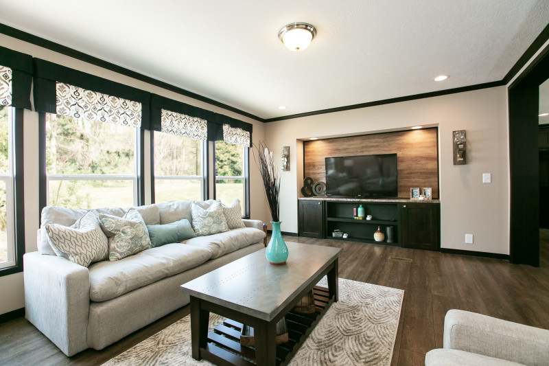 Living room of a manufactured home featuring four large windows, dark brown and beige furniture and decor and a recessed built-in entertainment center with storage and a wood accent wall.
