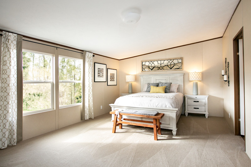 View is of a manufactured home bedroom with two large windows to the left. The room is neutral colors with a white bed and bed frame plus a wooden bench at the foot of the bed.