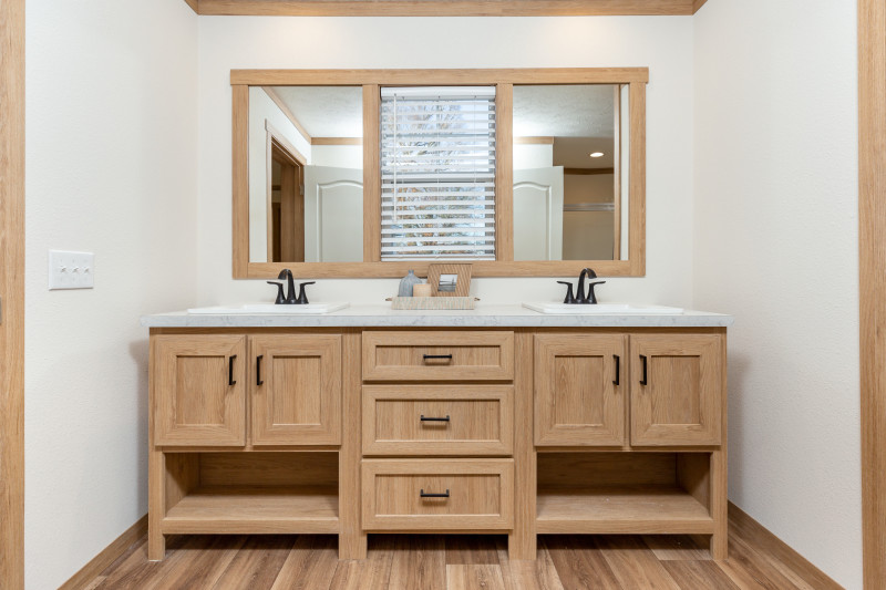 A primary bathroom double vanity. Two natural wood trimmed mirrors with a window in between and polished quartz-style counter tops.