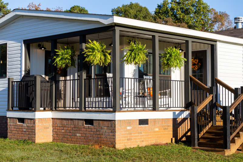 A manufactured home with a wraparound porch with black railing and beams and hanging plants.