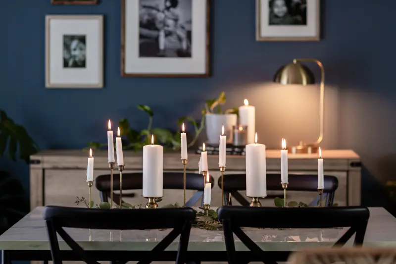 Row of white lit candles in gold holders sit on a table in a dining room with a light wood sideboard, black chairs, framed photos and a dark blue wall in the background.