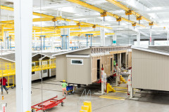 Inside of a manufactured home building facility with a white ceiling and concrete floor, with people working on three different home sections.