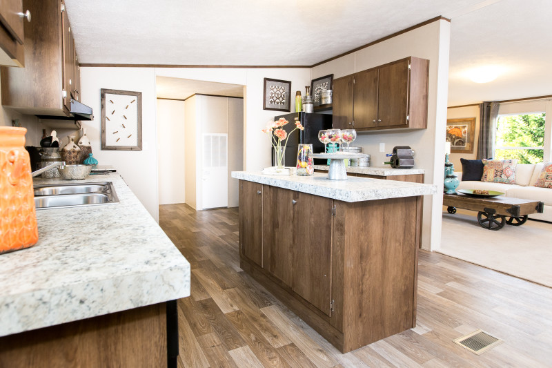 Kitchen of a manufactured home with brown cabinets, white counter tops and vinyl wood style flooring that opens into a living room. 